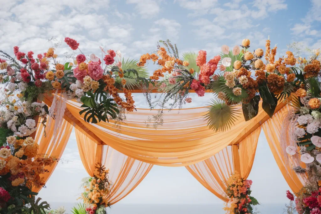 Orange and blush draped mandap edged with tropical florals, the ocean behind it