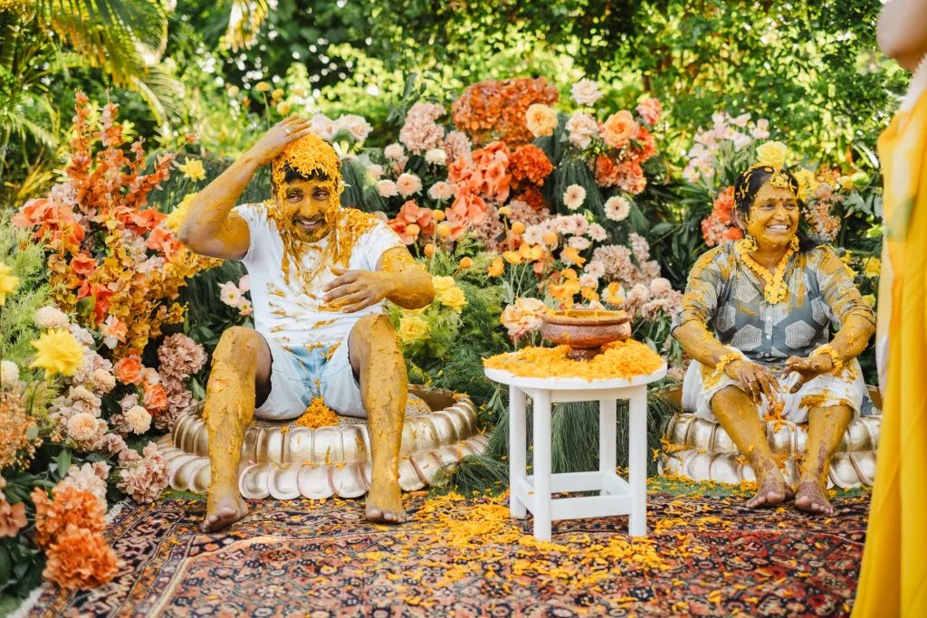 The couple laughing, covered in turmeric, seated on gold stools on a patterned rug in front of a wall of marigolds and orange blooms