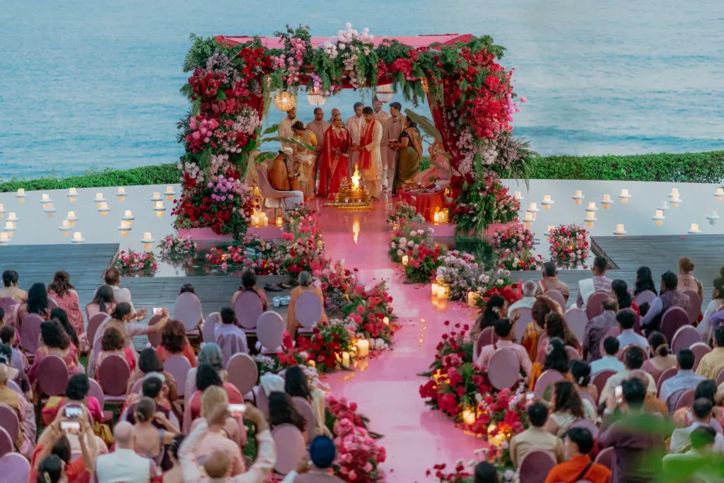 Pink and red floral mandap above the ocean at dusk, the couple at the fire with a candlelit aisle of petals running back through the seated guests
