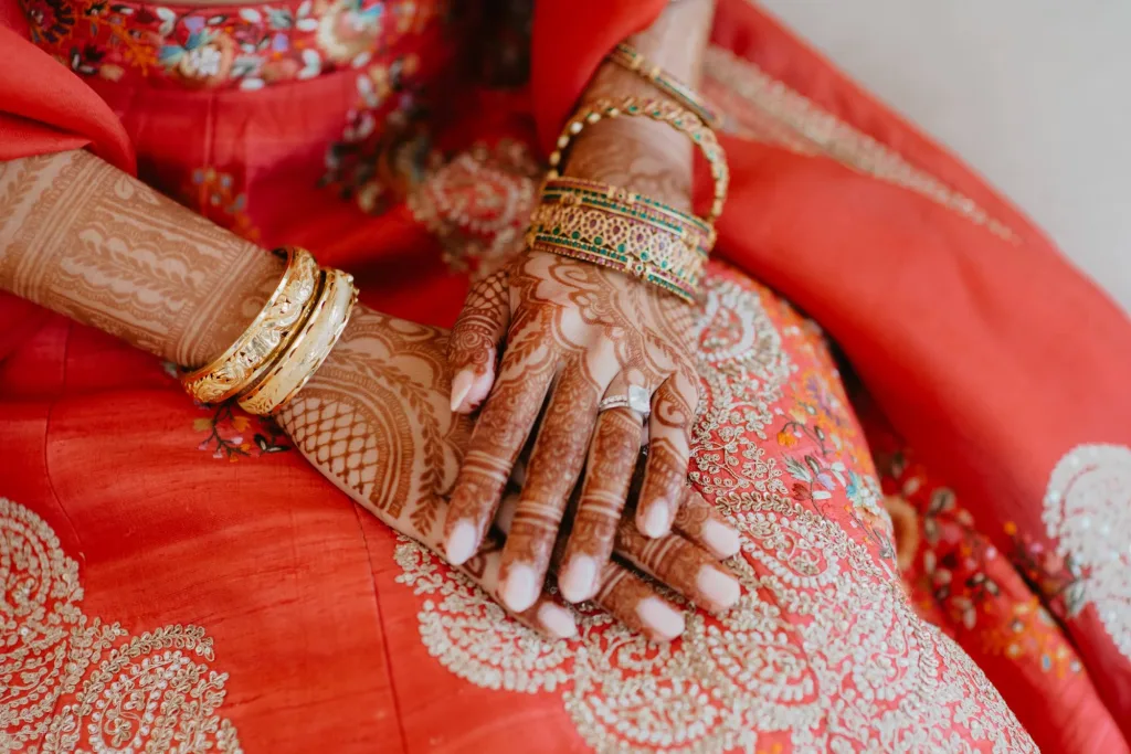 A bride’s hands resting in her red and gold lehenga, both palms covered in fresh mehndi and stacked with gold bangles