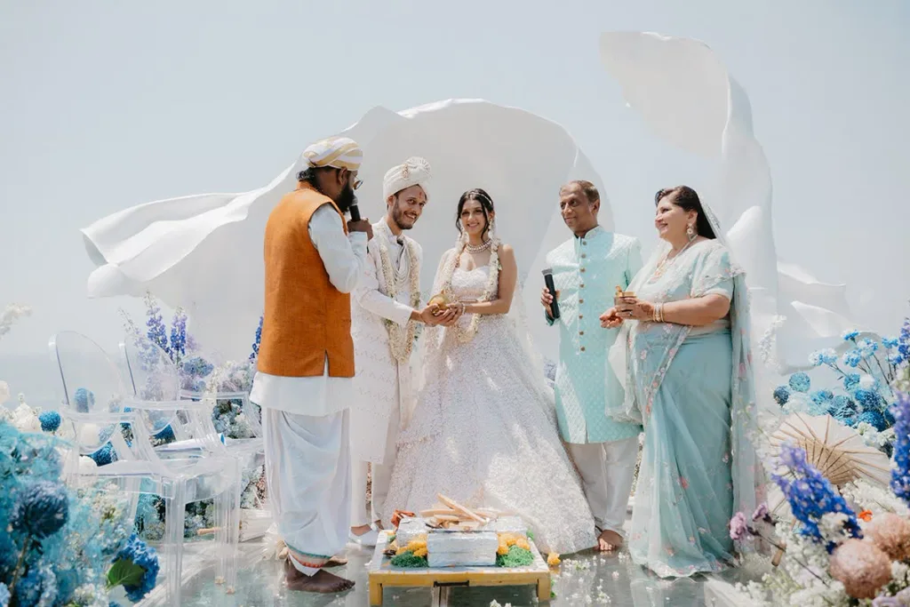 A priest leading the fire ceremony in front of a white backdrop and blue florals, the couple and their parents gathered around